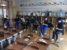 掲示物が並ぶ明るい教室内で、黒いTシャツや青いジャージを着用した6人の生徒たちが、椅子に座って譜面台の楽譜を真剣に見つめながらサックスを構え、パート練習やアンサンブルに集中して取り組んでいる風景の写真