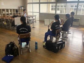 陽光が差し込む明るい教室内で、黒いTシャツや青いジャージを着用した4人の生徒たちが、椅子に座って譜面台の楽譜を真剣に見つめながらトランペットのパート練習や音出しに集中して取り組んでいる風景の写真