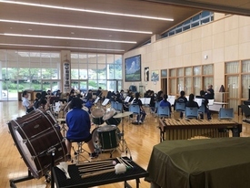 窓から陽光が差し込む広々とした音楽室内で、手前に大きなバスドラムやドラムセットなどの打楽器が配置され、その奥で大勢の吹奏楽部員たちが椅子に座って譜面台の楽譜を見ながら合奏練習に励んでいる活気ある風景の写真