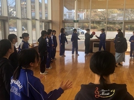 明るい屋内にて、青や黒のジャージを着た大勢の中学生が円状に並び、正面の二人が赤いボールを使いながらレクリエーションを行っている写真