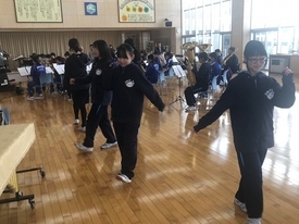 明るい室内にて、後方で吹奏楽部員たちが楽器の演奏練習に励む中、手前ではジャージを着た数名の生徒たちがリズムに合わせてステップを刻んだりポーズをとったりして、楽しそうにダンスの練習をしている様子の写真