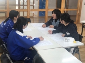 明るい室内にて、机を囲んで座る吹奏楽部員たちに対し、女性の指導者が手元の資料を指差しながら熱心に助言を行い、生徒たちが真剣な表情でその言葉に耳を傾けている写真