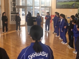 陽光が差し込む明るい室内で、お揃いの青いジャージや黒い服を着た中学生たちが輪になって集まり、中心にいる一人の生徒が赤いボールを手に持ちながら、周囲の仲間と笑顔でレクレーションを楽しんでいる写真