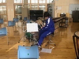 木目調の床が広がる明るい音楽室内で、青いジャージを着た一人の生徒がドラムセットの前に座り、譜面台に置かれた楽譜を真剣に見つめながらスティックを手に持ち、打楽器練習に取り組んでいる写真