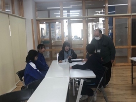 木目調の床が広がる明るい室内で、白いテーブルを囲んで座る中学生達と指導者が傍らに立ち、ミーティングを行っている写真