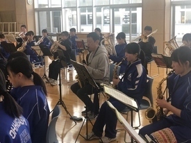 窓から陽光が差し込む明るい音楽室内で、大人の男性指導者が椅子に座り、青いジャージ姿の生徒たちと共に並んでサックスを演奏している写真