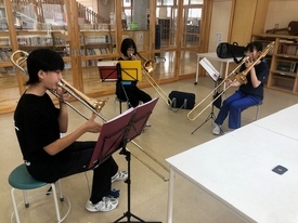 木目調の床が広がる明るい室内で、黒いTシャツや青いジャージを着用した3人の生徒が椅子に座って間隔を空けて並び、譜面台に置かれた楽譜を真剣に見つめながらトロンボーンのパート練習に励んでいる風景の写真