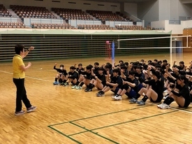 体育館で黄色のシャツを着た女性の先生が、身振り手振りで床に座る大勢の黒いウェアの生徒たちに向かって熱心に指導を行っている写真