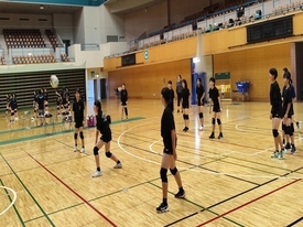 体育館のコート内で、黒いウェアを着た生徒たちがレシーブの低い姿勢で構えて練習に取り組んでいる写真