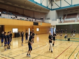 広い体育館のコート内で、黒いウェアを着用した生徒たちが飛んできたバレーボールを追いかけ、周囲の仲間や指導者がその動きを注視しながら適切なポジション取りやレシーブのタイミングを確認している写真