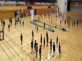広い体育館のコート内で、黒いウェアを着た大勢の生徒たちがネットを挟んで複数のグループに分かれ、練習している様子を上から撮影した写真