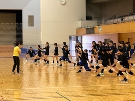 体育館にて黄色いシャツを着た先生の指示を受けながら、黒い練習着姿の大勢の女子生徒たちが、コート上で一斉に膝を深く曲げて片足を伸ばしている写真