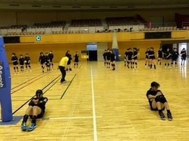 広い体育館のコート内で、黒い練習着を着用した多くの女子生徒たちが、手前で腹筋をしたり奥で整列し、先生の指導のもとで練習を行っている写真