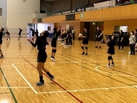 広い体育館のコート内で、黒い練習着を着用した多くの女子生徒たちが、ボールを使い練習を行っている写真