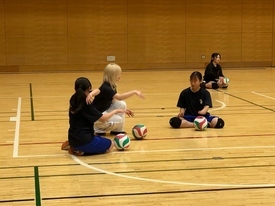 体育館のコート内で、バレーボールを前に床に座りながら、金髪の女性が身振りでアドバイスを送り、周囲の生徒たちが真剣な表情でその話を聞いている様子の写真