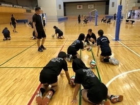 明るい体育館のコート内で黒い練習着を着用した複数の生徒たちが床に膝をついて、ボールを使った練習をしている写真