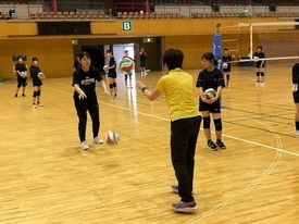 広い体育館のコート内で、黄色いシャツを着た先生が生徒に向かってバレーボールを投げ、黒い練習着の選手がそれを受け止める動作をしている写真