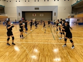 広い体育館のコート内で、黒いウェアを着た生徒たちが二列に向かい合って整列し、各自がバレーボールを顔の高さに構えてハンドパスを行っている写真