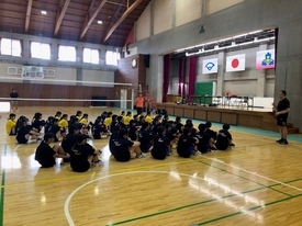 トレーニングウェアを着用した大勢の生徒たちが体育館の床に座り、ステージ前に立つ指導者の話に真剣な表情で聞き入っている写真