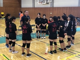 体育館のコートにて、黒い練習着を着用した女子生徒たちが指導者を半円状に囲み、真剣な表情で話を聞いている写真