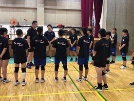 体育館のコートにて、黒いウェアを着用した女子生徒たちが、男性指導者を囲んで半円状に並び、話を聞いている写真