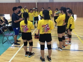 体育館にて、黄色いTシャツを着用した女子生徒たちが指導者を囲んで円になり、真剣な表情で話を聞いているミーティング風景の写真