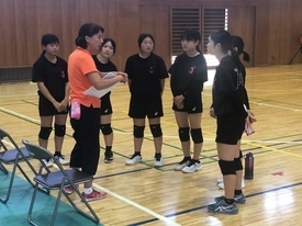 体育館にて、オレンジ色のポロシャツを着た女性指導者がバインダーを手に持ちながら、黒い練習着を着用した数名の女子生徒たちに近距離で熱心にアドバイスを送っているミーティング風景を捉えた写真