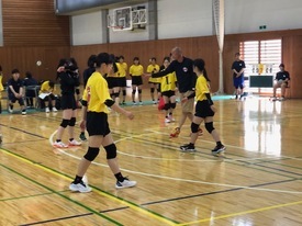 体育館のコートにて、黄色いTシャツを着用した女子バレーボール選手たちが、指導者から動作のアドバイスを受けている写真