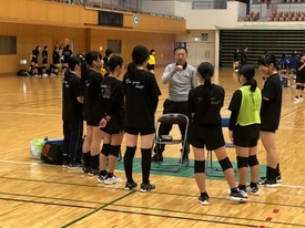 体育館にて、黒いトレーニングウェアを着用した女子生徒たちが先生を囲み、真剣な表情で話を聞いている写真