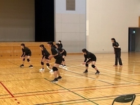 広い体育館にて、黒いウェアを着用した5人の女子生徒たちが、低い姿勢でレシーブの構えを取っている写真