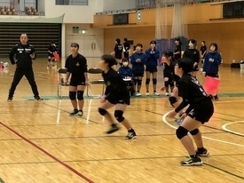 体育館のコートにて、黒いユニフォームを着用した女子生徒たちが、低い姿勢でレシーブの構えを取りながら、真剣な表情でボールの行方を追っている写真