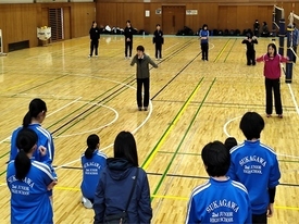 体育館で青いジャージを着た生徒たちが手前に並び、コート中央に立った2名の先生が手を後ろに伸ばし指導を行っている練習風景を捉えた写真