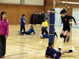 体育館で、床に座った仲間の足を飛び越えるトレーニングに励む女子生徒たちの姿を、ピンク色のウェアを着た先生が見守っている様子の写真