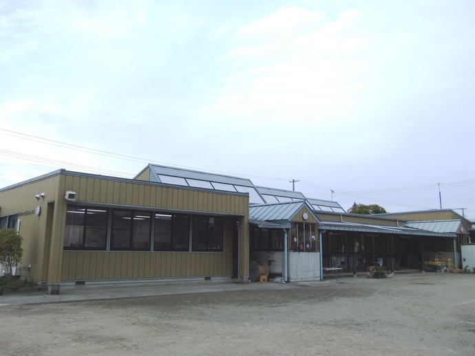 薄い黄土色の縦縞の外壁と、天窓が並ぶ特徴的なグレーの屋根を持つ平屋建ての園舎が、広く平坦な砂地の園庭に面して横に長く伸びており、曇り空の下で落ち着いた雰囲気を見せている、モダンな造りの旧須賀川市立長沼幼稚園の写真