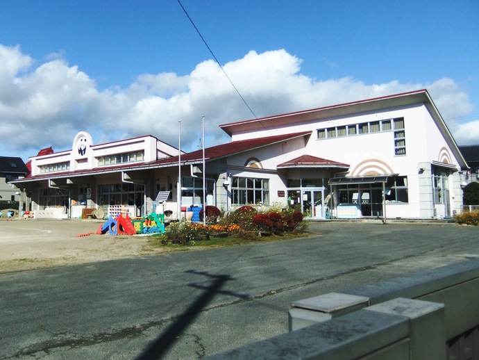 青い空と白い雲の下、赤い屋根と白い外壁が特徴的な平屋建ての旧須賀川市立仁井田幼稚園の園舎が建っており、園庭には色鮮やかな遊具や花壇が配置され、左側の建物上部にはパンダのマークが掲げられている、日当たりの良い園庭と建物の写真