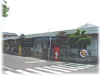 道路に面して建つ白い平屋建ての建物の外観で、手前には横断歩道があり、門門付近には赤い看板や可愛らしいキャラクターの円形パネルが設置された、親しみやすい雰囲気の保育施設を斜めから撮影した写真