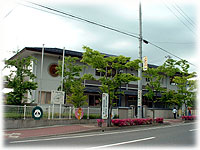 街路樹の緑や色鮮やかなピンクの花に囲まれた、落ち着いたグレーの外壁を持つ二階建ての第二保育所外観写真