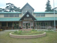 三角形の大きな屋根と中央に配置された時計が特徴的な、木材を活かした温かみのある平屋建ての「白方こども園」の正面玄関を、手前に円形の花壇が広がる芝生エリアから捉えた写真