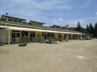 青空の下、背後には緑豊かな木々が広がり白い外壁にクリーム色の大きな日除けタープが複数設置されて砂地の広い庭に面して建つ平屋建ての施設の写真