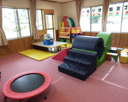 明るい光が差し込む室内に、赤色の円形トランポリンや青色の階段状ソフト遊具、滑り台などが配置された、子どもたちが安全に体を動かして遊べるよう配慮された広々としたプレイルームの様子を捉えた写真