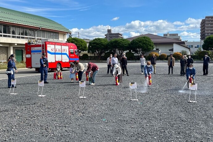 広い屋外で消防車を背に複数の参加者が並んで消火器訓練を行い白い噴霧を放つ様子の写真