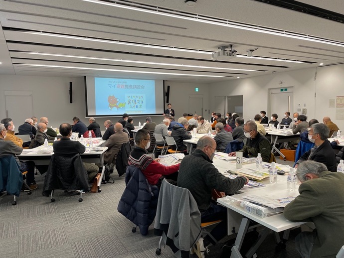 広い会議室で多くの参加者がグループに分かれて着席し、前方スクリーンの前に立った男性の話を聞いている講習会の様子の写真