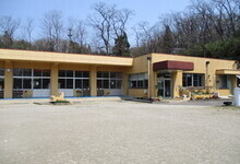 平屋建てのベージュ色の校舎と、その手前に広がる静かな土のグラウンドを、晴天の下で遠くから捉えた、稲田幼稚園の全景の写真