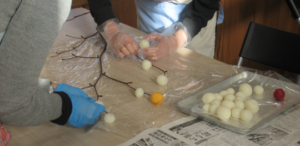 ビニール手袋やゴム手袋をはめた参加者が、トレイに用意された白や黄色、赤色の団子を、テーブルの上に置いた細い木の枝先に一つずつ丁寧に取り付けて飾り付けを行っている作業風景の写真