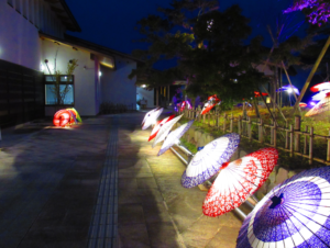 夜の通路に沿って、色鮮やかな和柄が設置された数多くの和傘が内側からライトに照らされて美しく並び、その幻想的な光が石畳の地面や周囲の木々を照らし出している風景を捉えた写真