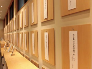 短冊形の作品が掛かった木製パネルが、整然と壁一面に並んだ展示の様子の写真