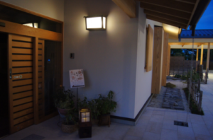夕暮れの和風の玄関先に柔らかな灯りがともり、植木鉢や小さな灯籠が並ぶ静かな通路が奥へ続く様子の写真