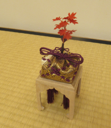 畳の上に置かれた小さな白木の台座に、金糸が織り込まれた紫色の袋が載り、その上部に鮮やかに紅葉したモミジの小枝が添えられた床飾りの写真