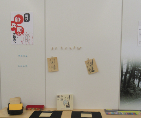 白い壁面に「卯兵衛さんを倣おう」と書かれたチラシと、筆絵風の絵が描かれた2枚のカードが貼られ、下の台にはメモ用紙などが置かれている写真