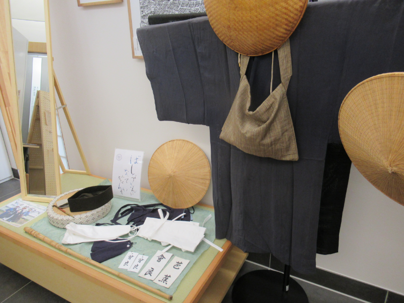 濃い灰色の旅装束と編み笠、布袋や帯など昔の旅道具が並ぶ展示の写真